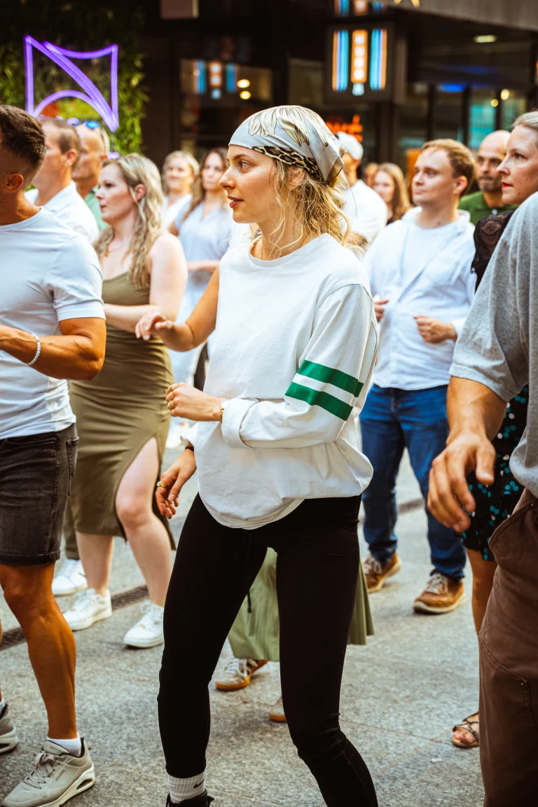 A woman wearing a headscarf dances outdoors in a crowd under natural daylight, surrounded by other people.