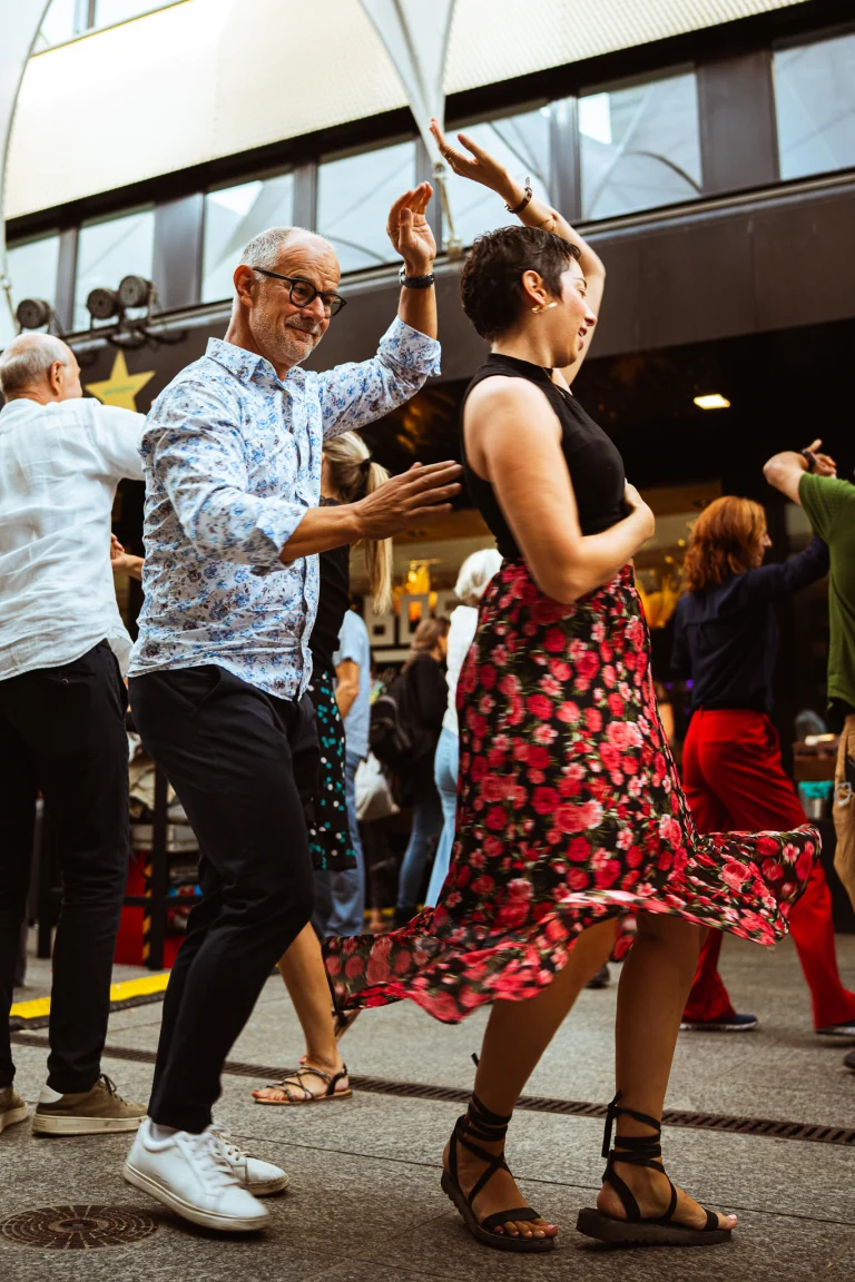 A man and a woman dance together outdoors on a paved ground, surrounded by other dancing couples.