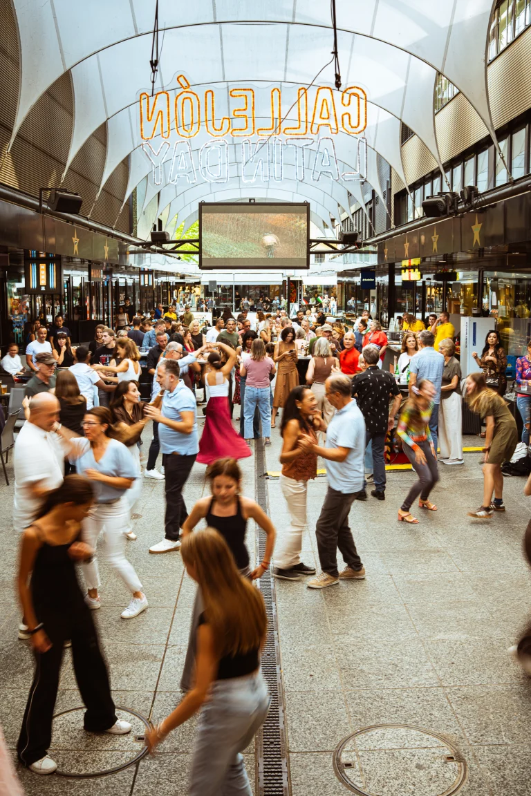 A large crowd of men and women dance in a covered, arched pedestrian street beneath a glowing sign reading CALLEJÓN LATIN DAY.