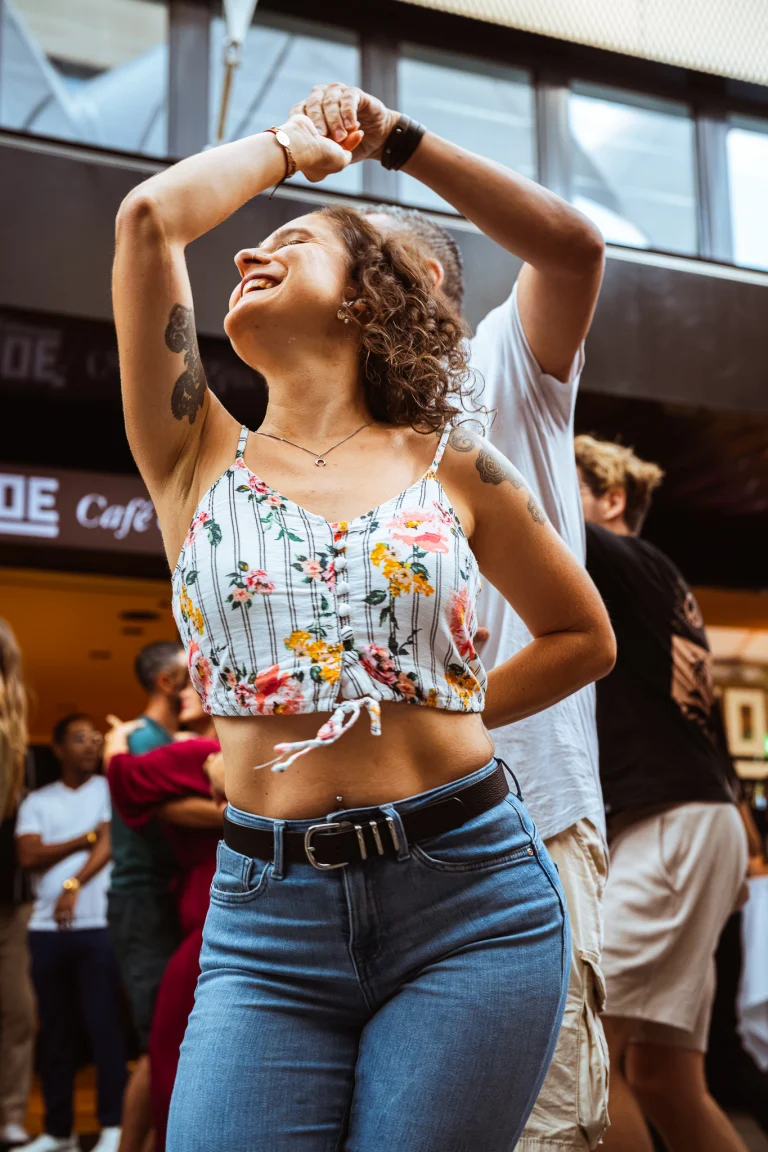A smiling woman with curly hair and a floral crop top dances, holding hands with a man behind her.