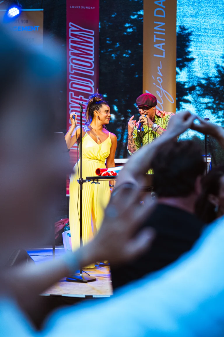 A woman in a yellow jumpsuit and a man in a patterned shirt singing into microphones on stage, viewed past blurred audience members.