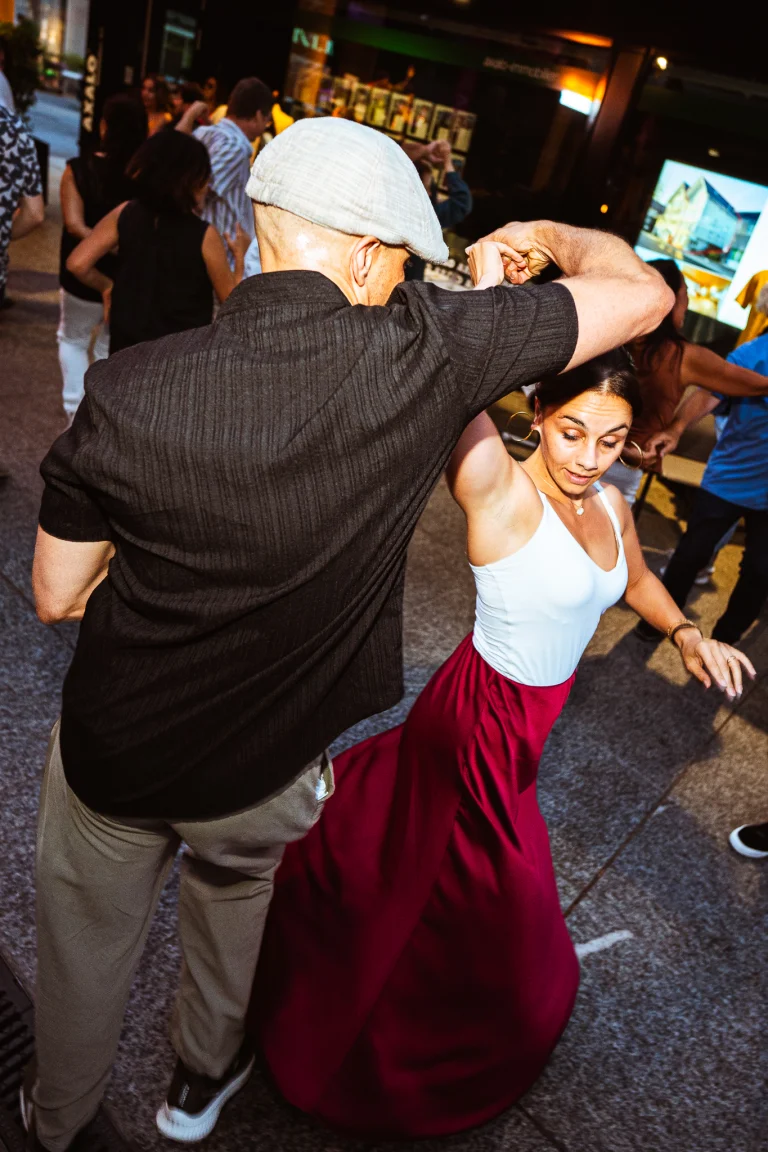 A man in a cap and black shirt dances with a woman in a white top and red skirt on an outdoor pavement.