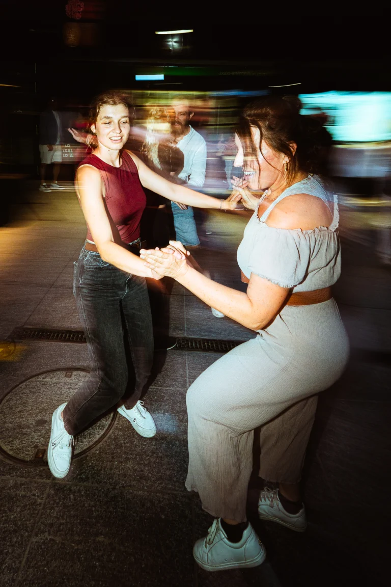 A woman and a woman dance and hold hands at night, captured with motion blur and direct flash lighting.