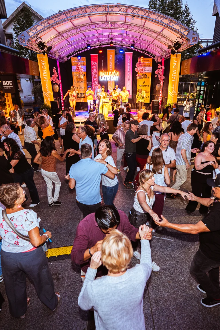 Couples dance on an outdoor plaza beneath a lit stage with banners reading Callejón Latin Day.