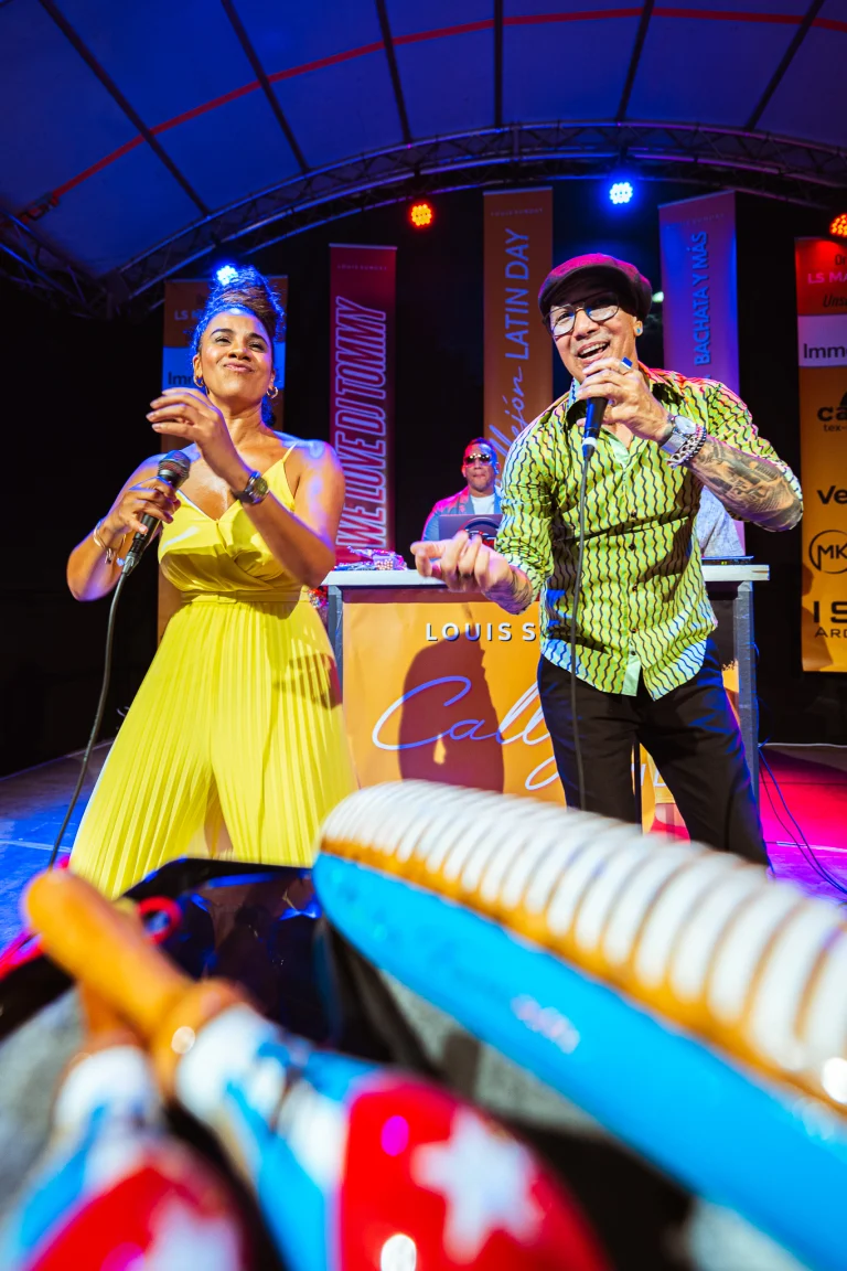 A woman in a yellow dress and a man in a patterned shirt sing into microphones on a stage with percussion instruments in the foreground.