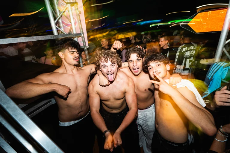 Four shirtless young men pose closely together at night under motion-blurred colored lights, with one sticking out his tongue and another making a peace sign.