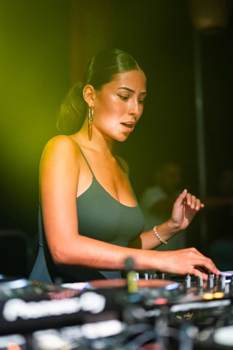 A woman stands behind a DJ mixer under green and warm stage lighting, operating the controls.