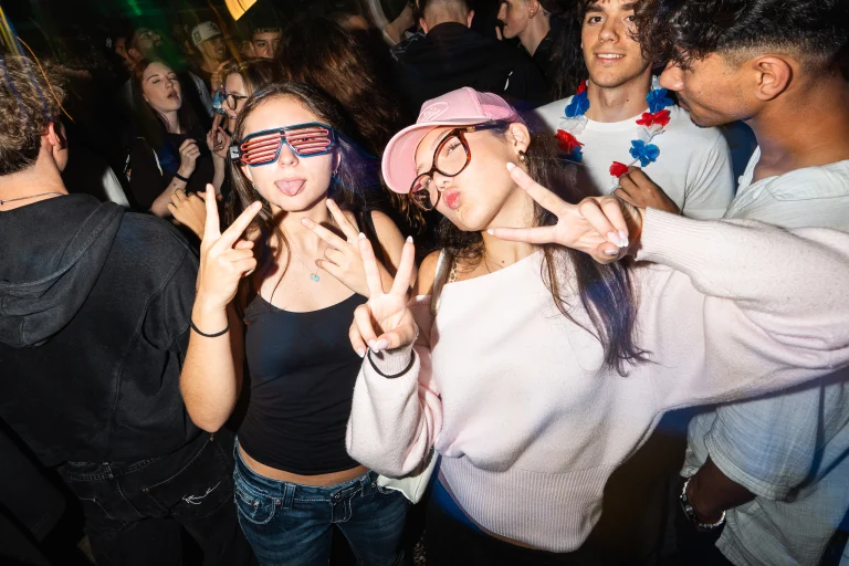 Two young women flash peace signs in a crowded venue, with one wearing shutter shades and sticking out her tongue, and the other wearing a pink cap while puckering her lips.