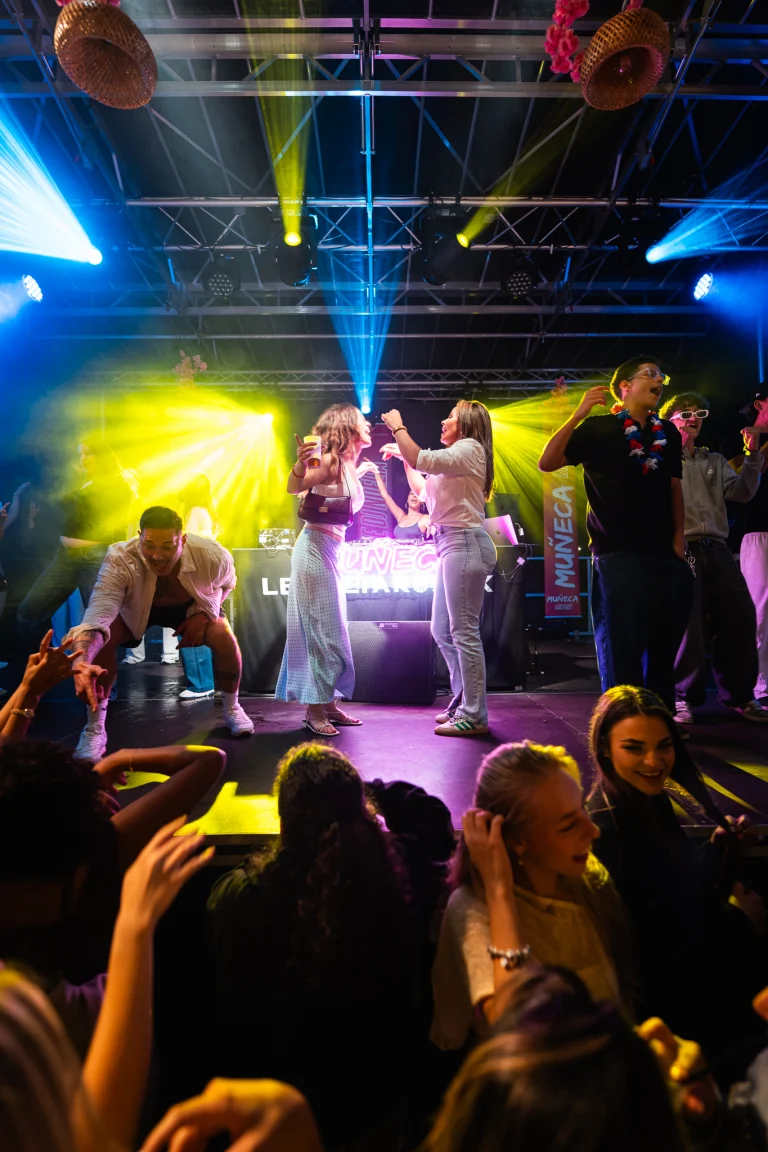 People dance and celebrate under colorful stage lights at an outdoor evening festival.