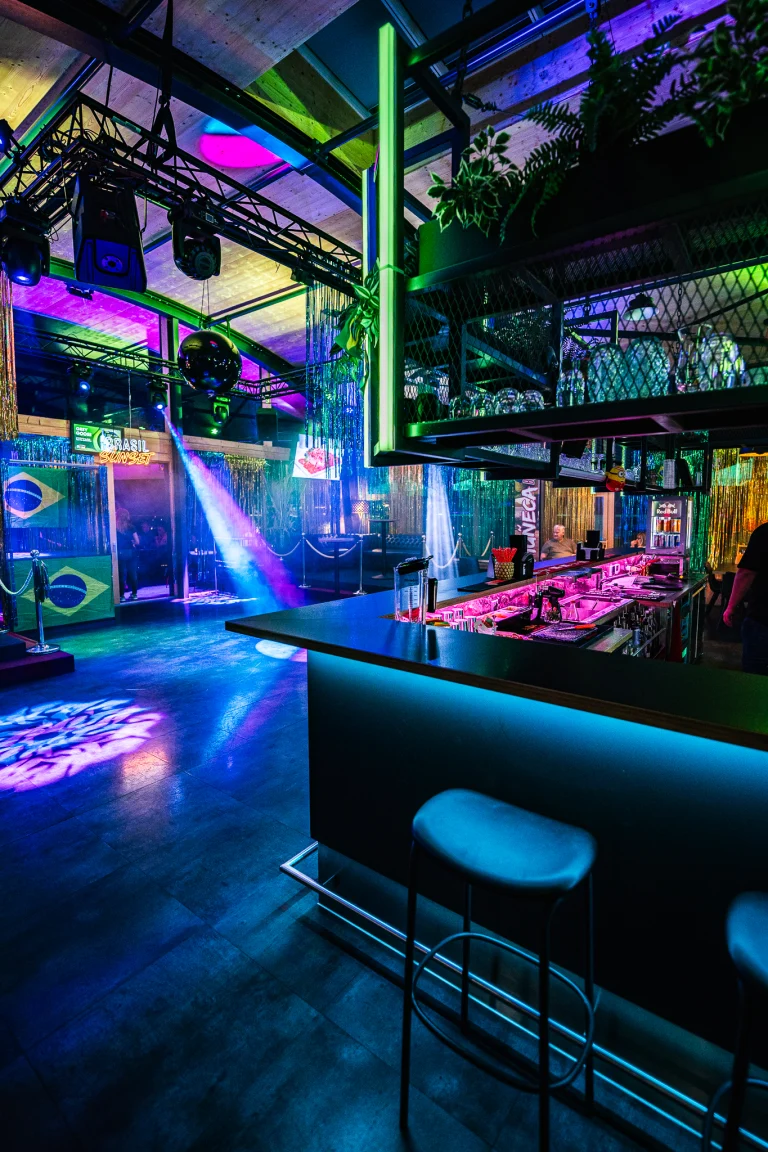 An indoor venue with a bar lit by blue and purple lights, featuring stools, a dance floor, and Brazilian flags.