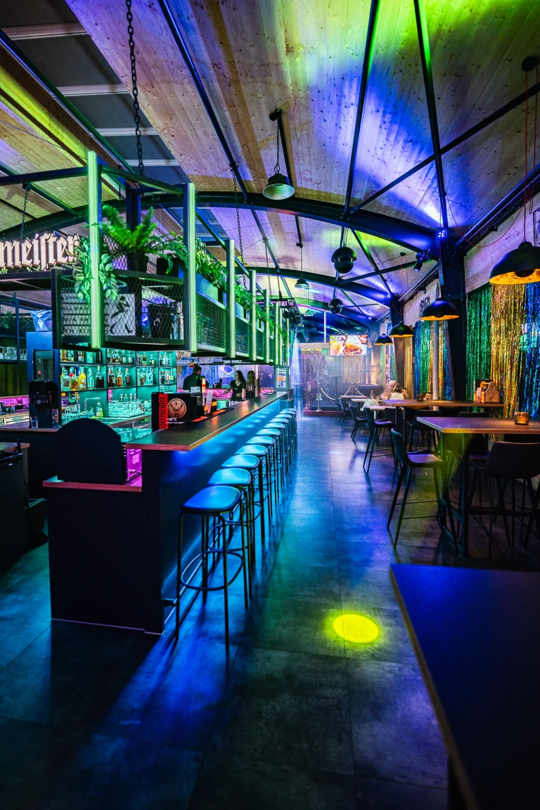 An indoor bar illuminated by blue and green lighting, featuring a long counter with high stools on the left and tables on the right beneath a metal truss ceiling.