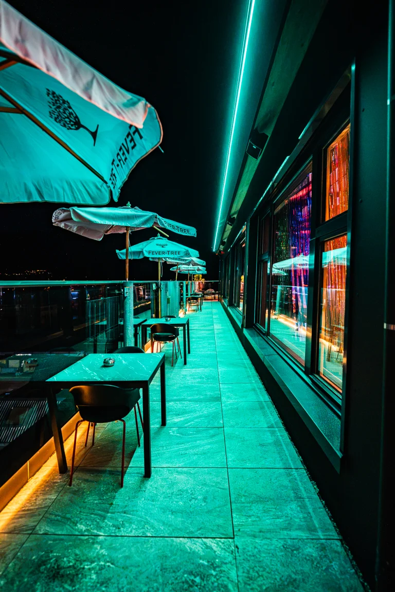 An empty rooftop terrace at night illuminated by green lighting, featuring tables, chairs, and parasols along a building facade.