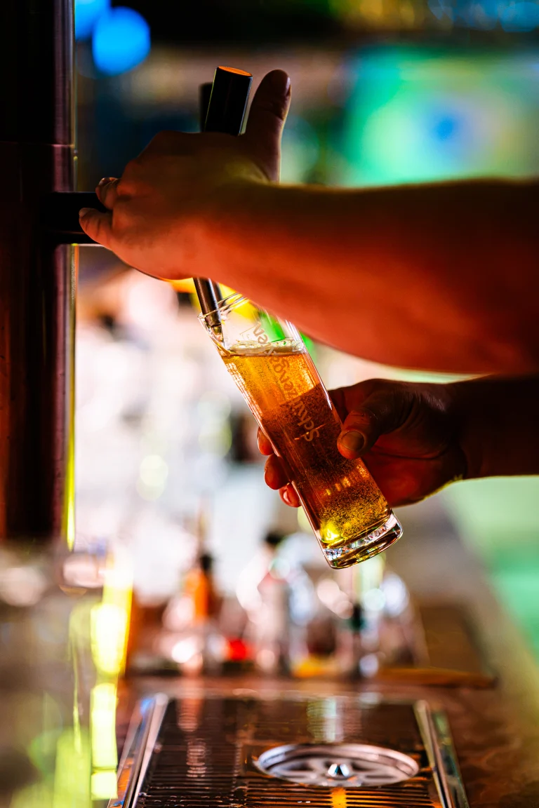 A person pours golden beer from a tap into a tall glass at a bar.