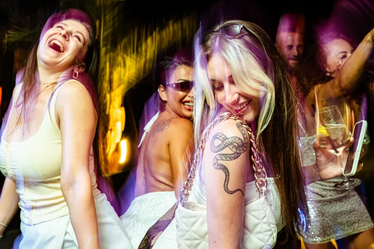 Several smiling women dancing at night under direct flash lighting, with one woman in the foreground displaying a prominent snake tattoo on her shoulder.