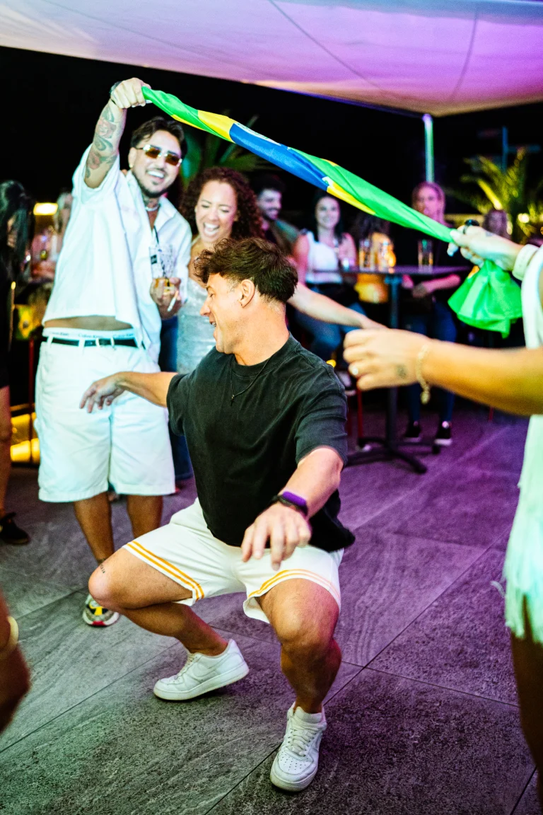 A man in a dark shirt and white shorts dances in a low squat, while people in the background hold a green, yellow, and blue flag.