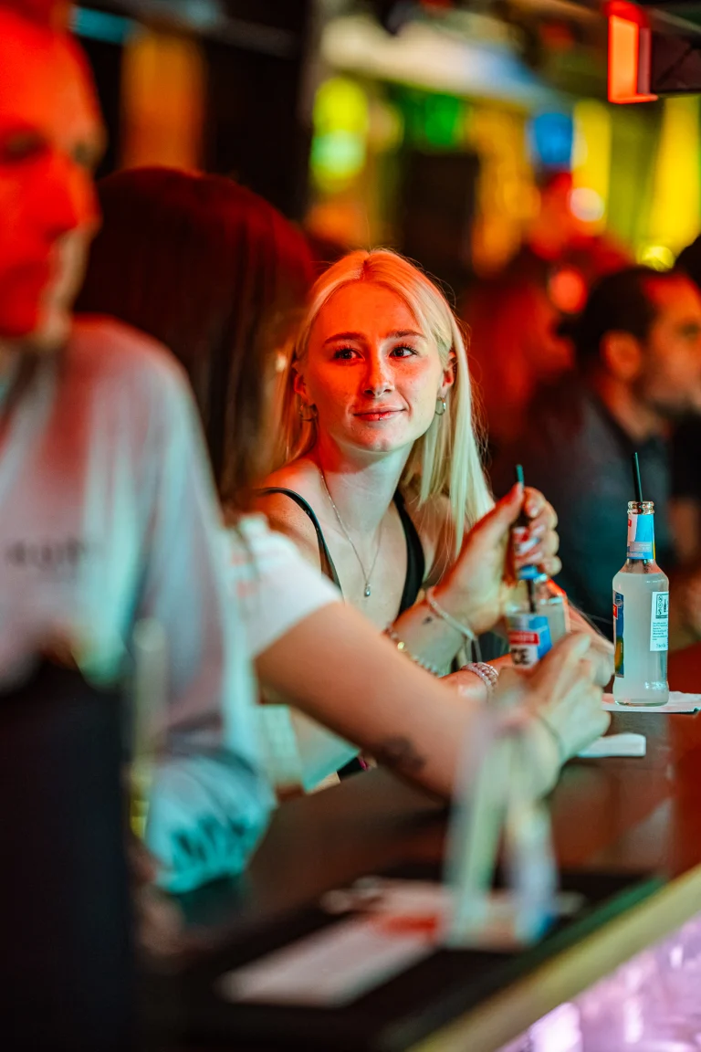 A blonde woman sits at a bar under warm indoor lighting, holding a bottle with a straw while people sit nearby.