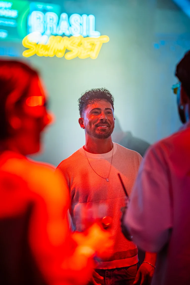 A smiling man in a white knitted shirt stands indoors under red lighting, with silhouettes of other people in the foreground and a neon sign reading BRASIL SUNSET in the background.