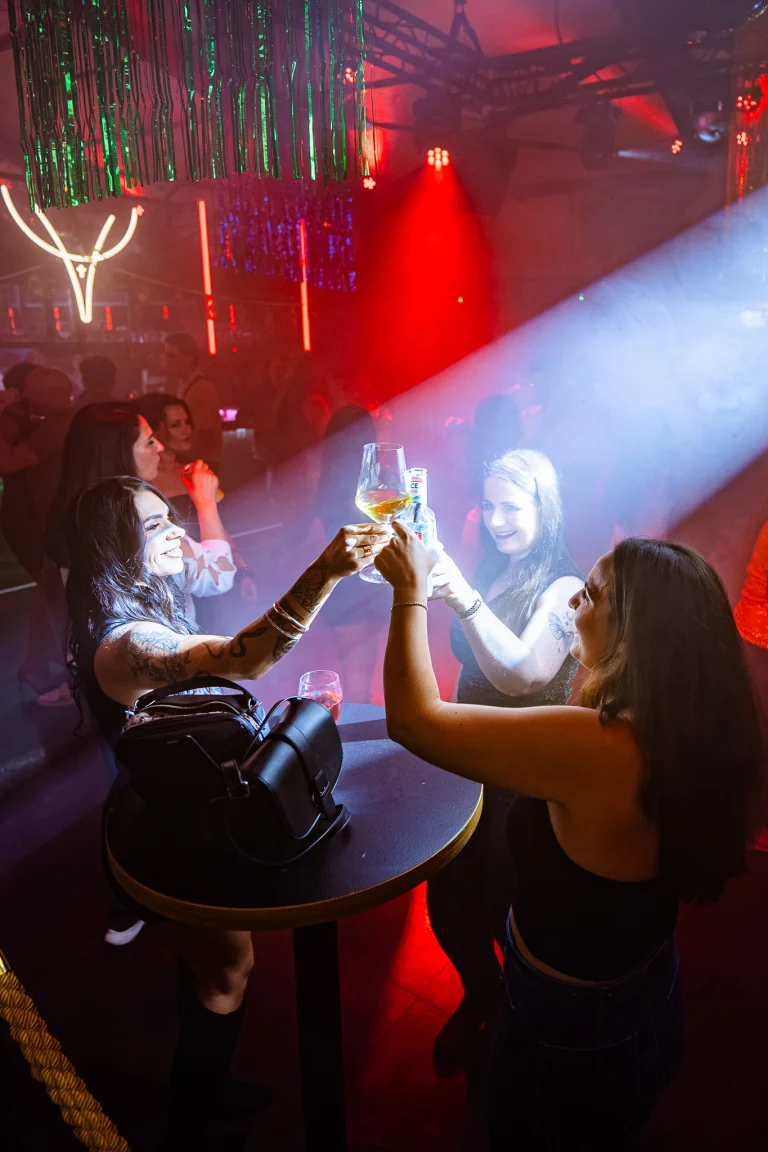 Three women in a dimly lit club raise their glasses in a toast over a high round table, illuminated by a beam of blue light and red ambient glow.