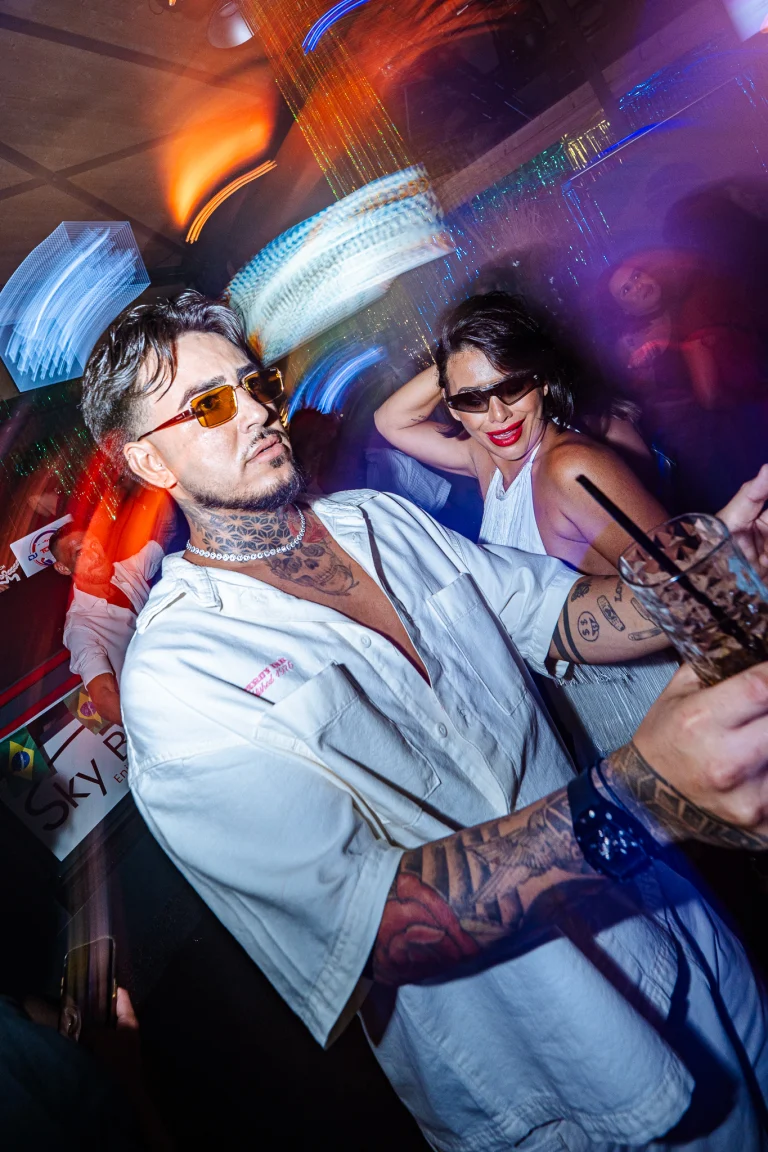 A tattooed man in a white shirt and sunglasses holds a drink in a dimly lit, colorful nightclub, while a woman in a white top dances in the background.