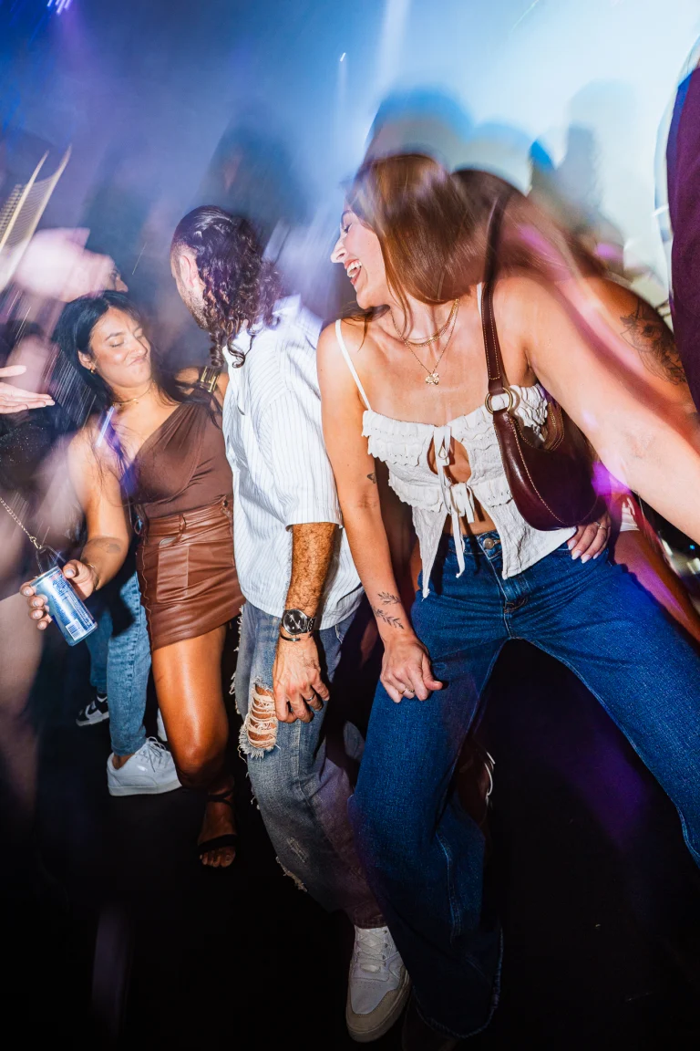 A wide-angle, motion-blurred shot of a woman in a white top and jeans dancing in a crowd with other people.