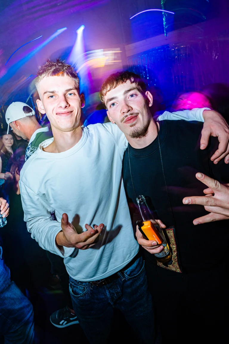 Two young men pose together in a crowded nightclub with blue and purple overhead lighting, one holding a glass bottle.